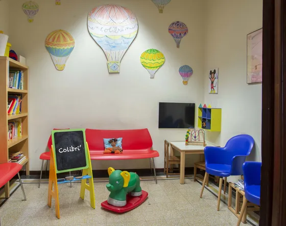 Sala d’attesa con mongolfiere dipinte sulle pareti, divano rosso e lavagnetta con la scritta Colibrì