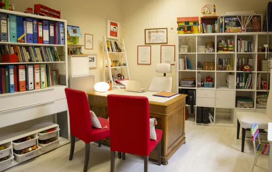 Stanza di psicopedagogia con scrivania, sedie rosse e scaffali di libri e giochi
